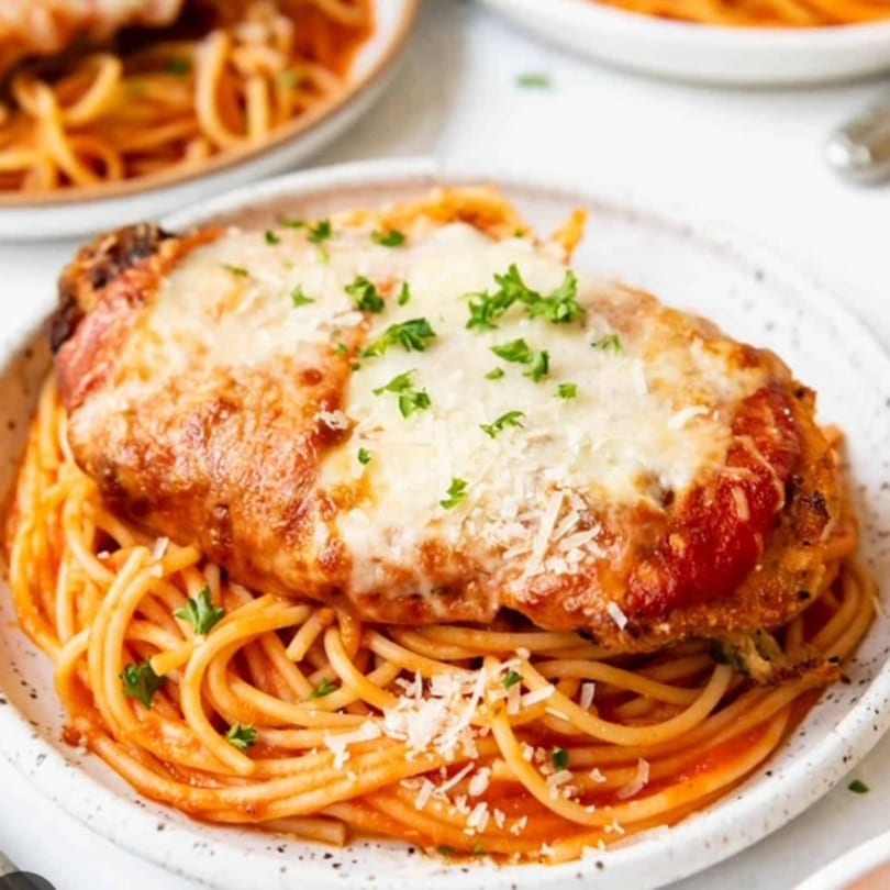 Chicken parmesan with melted cheese and red sauce, served over spaghetti on a white plate.